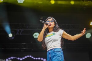 Female singer on stage wearing sunglasses, holding a microphone and singing passionately.