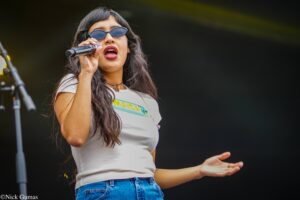 Female singer on stage wearing sunglasses, white t-shirt and jeans, singing into a microphone.