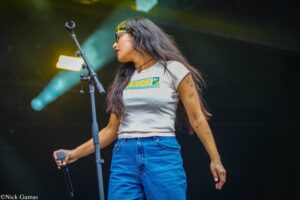 Female singer on a concert stage, holding a microphone, wearing a white Jamaica graphic t‑shirt and blue jeans.
