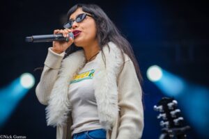 Female singer performing on stage, holding a microphone near her mouth, wearing sunglasses and a white fur vest.