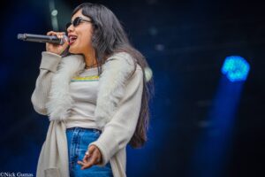 Female singer wearing sunglasses, white top and fur vest, holding a microphone mid-performance on stage.