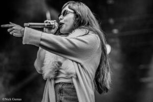 Female singer on stage, wearing sunglasses, singing into a microphone while pointing with her left hand.