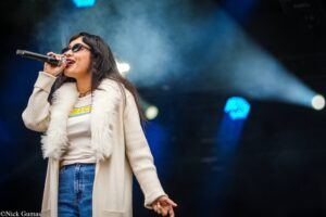 Female singer on stage wearing sunglasses and a beige cardigan, holding a microphone and singing into the crowd.