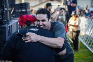 Two men share a warm hug at an outdoor event, one smiling broadly, with stage equipment and a blurred crowd in the background.