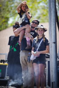 A group of people at an outdoor festival; a man lifts a child on his shoulders while others watch, smiling near a metal pole.