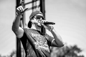 Male singer performing on an outdoor stage, wearing a backward cap and sunglasses, mid‑song with mic in hand and one finger raised.