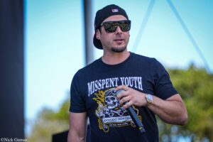 Man wearing a black cap and sunglasses, holding a microphone outdoors during a performance, with a graphic tee reading 'Misspent Youth'.