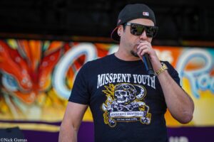Male vocalist in a backwards cap and sunglasses holding a mic, performing in front of bright graffiti art.