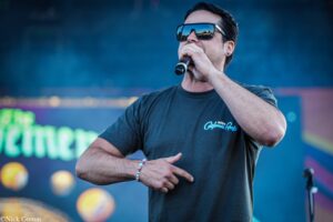 Male singer wearing sunglasses performs into a handheld microphone on an outdoor stage.
