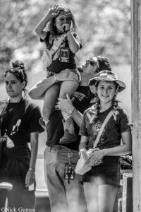 Man lifts a young girl onto his shoulders at an outdoor event as she waves to the camera.