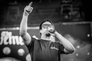 Male singer on stage wearing sunglasses, microphone in hand, raising his right fist with index finger pointed upward.