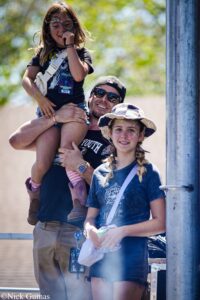 Man wearing a cap and sunglasses lifts a smiling girl onto his shoulders at an outdoor event, with another braided girl beside them and face paint visible on both.