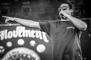 Male singer on stage with a microphone, wearing sunglasses, gesturing with his arm extended to the left toward the crowd.