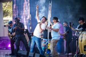 Outdoor concert stage with two vocalists at center; one singer raises his arm while the other holds a long decorative banner, smoke and band equipment around him.