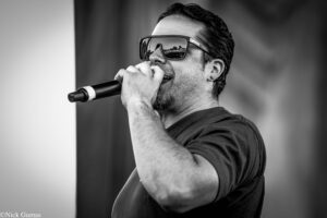 Male singer wearing sunglasses, close-up profile, singing into a microphone on stage (BW image).