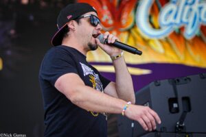 Male singer wearing a black cap and sunglasses performs into a microphone on a stage with a colorful graffiti backdrop behind him.