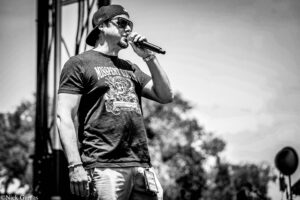 Male singer wearing a backward cap and sunglasses, holding a microphone on an outdoor stage.