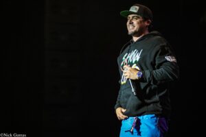 Male performer on a dark stage, holding a microphone and wearing a black hoodie and cap.