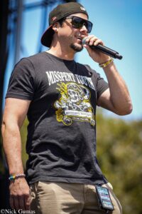 Male singer wearing sunglasses and a backward cap, holding a microphone on an outdoor stage, in a black "Misspent Youth" t-shirt.