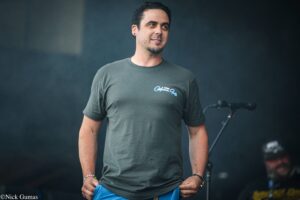 Man in a gray t-shirt on stage, looking to the right, hands at his waist with a blue cloth in front of him. The shirt shows a small white logo.