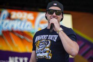 Male singer on stage wearing a black cap and dark sunglasses, microphone in hand, colorful graffiti backdrop behind him.