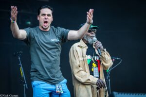 Two men on stage, one shouting with arms raised, the other holding a microphone and singing or speaking, dark backdrop behind them.