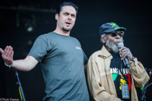 Two men on a stage; one in a gray shirt speaks while the other wears a Jamaica flag cap and sunglasses and holds a microphone.