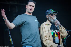 Two men perform on a dimly lit stage: a younger man in a gray t-shirt raises his left arm while singing, and an older man with a gray beard wearing a Jamaica-themed shirt, tan jacket, sunglasses, and a cap holds a microphone nearby.