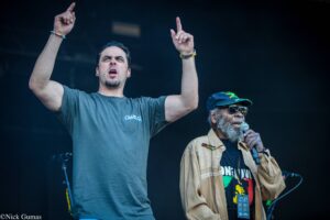 Two men on a dark stage: the foreground singer in a gray T‑shirt raises both arms and points upward, while the second man with a cap and sunglasses holds a microphone nearby.