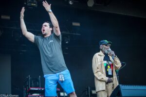 Two men perform on a stage: one raises arms in a gray shirt and blue shorts, the other sings into a microphone while wearing sunglasses and a beige jacket.