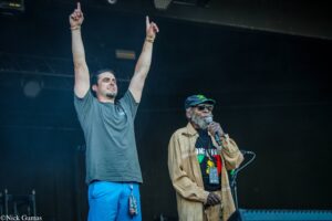 Two men on a dark stage: one in a gray shirt raises both arms with index fingers pointing up, beside another man wearing a cap and sunglasses holding a microphone.