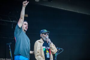 Two men perform on a dark stage; one raises his fist while the other speaks into a microphone.