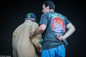Two men on a dark stage viewed from behind; one in a gray graphic T-shirt holding a microphone.