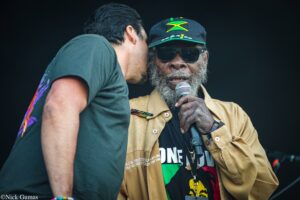 Two men on stage, one speaking into a microphone while the other leans in, wearing sunglasses and a Jamaican flag hat against a dark background.