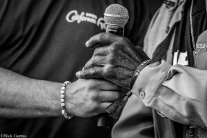 Close-up of a person gripping a handheld microphone during a performance, with folded arms and a beaded bracelet visible.