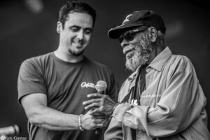 Two men on stage, one presenting a microphone to the other while they smile and talk back and forth on stage.
