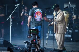 Back view of a musician wearing a gray t-shirt with a colorful 'California Roots' design on stage, guitar in hand and microphone stands nearby.