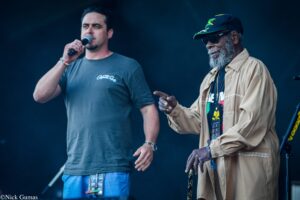 Two men on a dark stage; one sings into a handheld microphone while the other wears a cap and sunglasses.