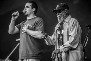 Two male performers singing on stage: one holds a microphone and has a hand on his chest, the other wears a cap and sunglasses with a gray beard, on a dark background (black and white).