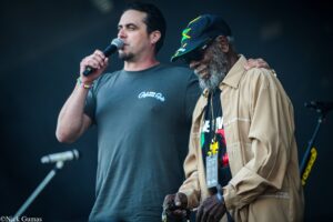 Two men on stage; one singer holds a microphone while the other, wearing a Jamaica-cap, smiles and leans in.