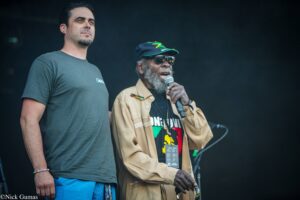 Two men on stage: a younger man in a grey t-shirt and blue shorts standing beside an older man in a tan jacket, cap, and sunglasses holding a microphone.