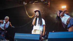 Three performers on a concert stage: center singer with microphone and braids, caps, and glasses, flanked by two dancers in shiny outfits under stage lights.