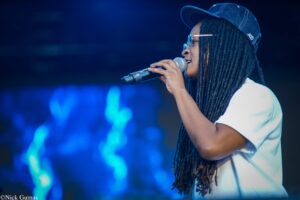 Singer wearing a denim cap and glasses, singing into a handheld microphone on a blue-lit stage (side profile).