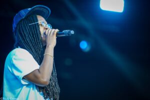 Singer with long dreadlocks wearing a blue cap and glasses, holding a microphone on a dimly lit stage.