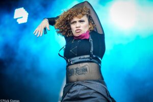 Female performer on stage with curly hair, in a black outfit with harness, raising her left arm under bright blue lights.