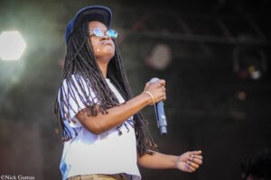 Singer with long dreadlocks wearing a blue cap and round sunglasses, holding a microphone on stage.