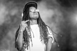 Female performer with dreadlocks, wearing a cap and round sunglasses, singing into a microphone on stage.