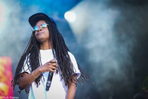 Female performer with long dreadlocks, wearing a cap and round sunglasses, holding a microphone on stage.