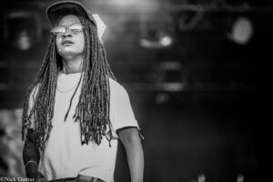 Black-and-white photo of a performer with long dreadlocks, a cap, and round sunglasses on stage, holding a microphone.