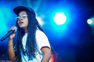 Female singer with long dreadlocks and glasses, holding a microphone on stage under blue lights.